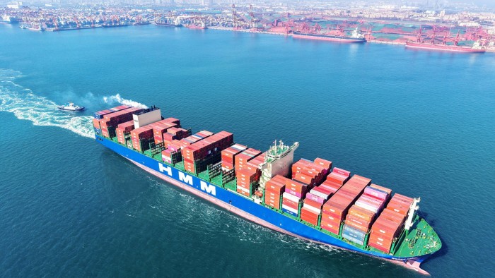 A large container ship is sailing through the blue waters of Qingdao port in China's eastern Shandong province. The vessel is loaded with numerous stacked shipping containers