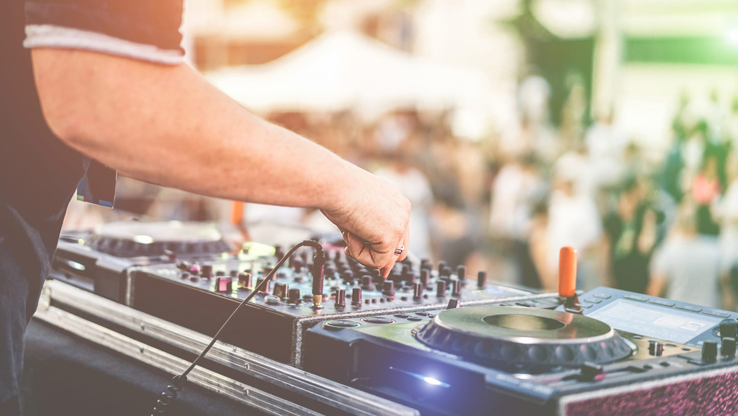 A close-up of hands at a DJ table with a blurred summertime crowd in the background