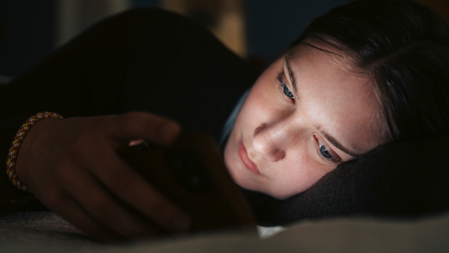 A teenage girl lies on her side in the dark, her face illuminated by her smartphone, suggesting she is interacting with something on the screen.
