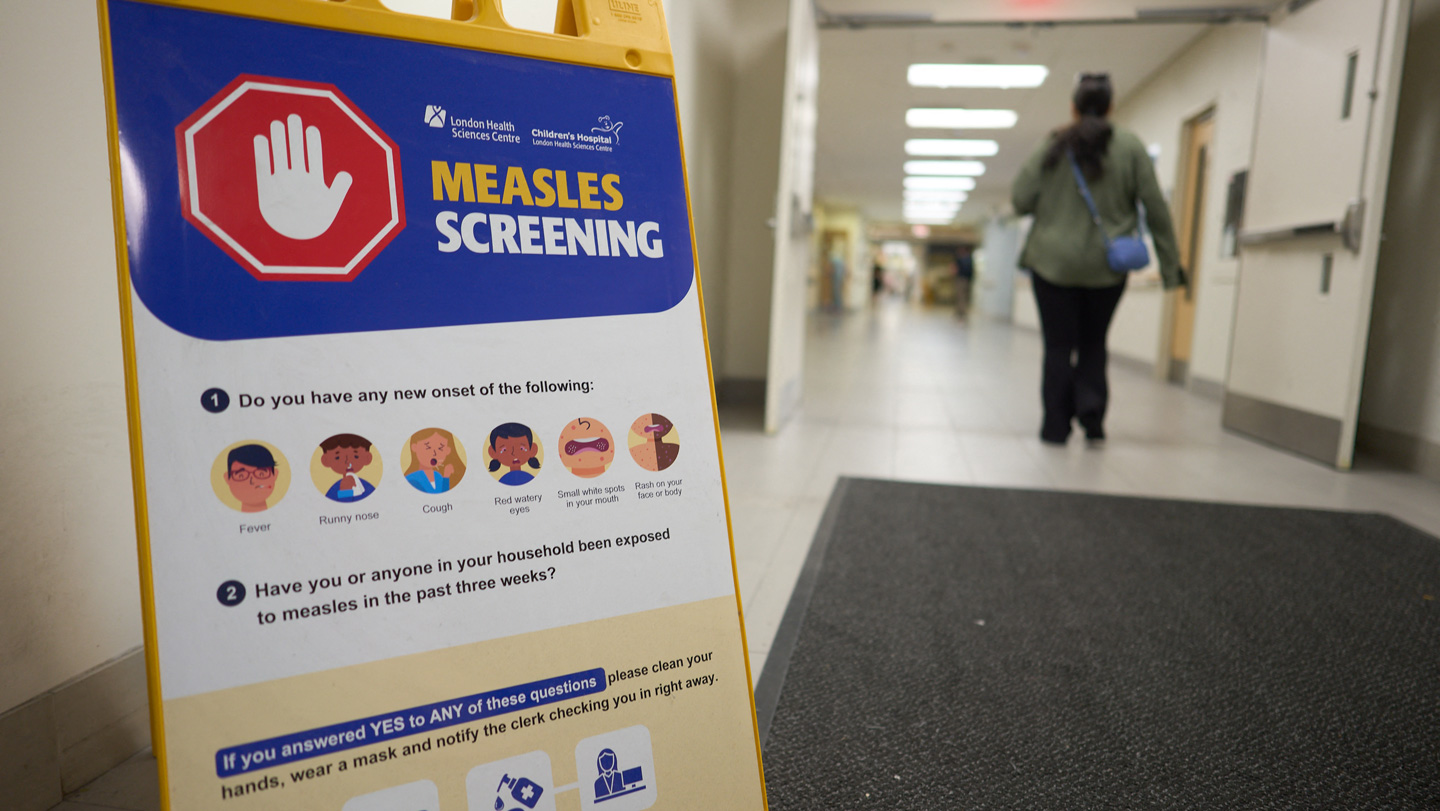 A sign advertising measles screening is in the foreground, to the left. The top of the sign has a red octagon with a white hand signaling stop, with "measles screening" written to the right. Underneath the sign lists measles symptoms and what to do if exposed to measles. To the right of a sign, a woman walks through open double doors into a hospital corridor.
